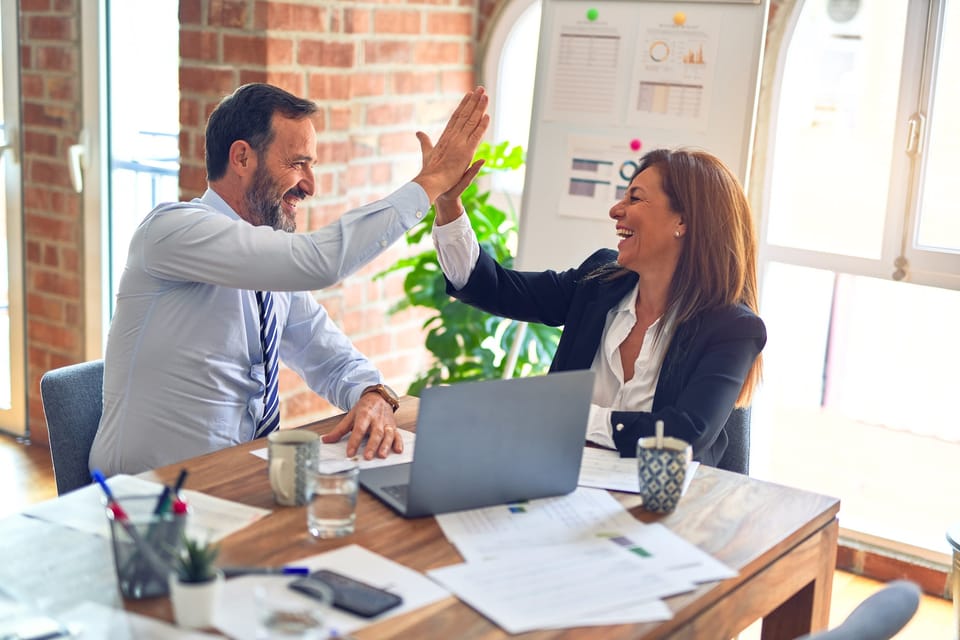 A man and a woman in suits high fiving each other while sitting at a cluttered table with business documents and a laptop.